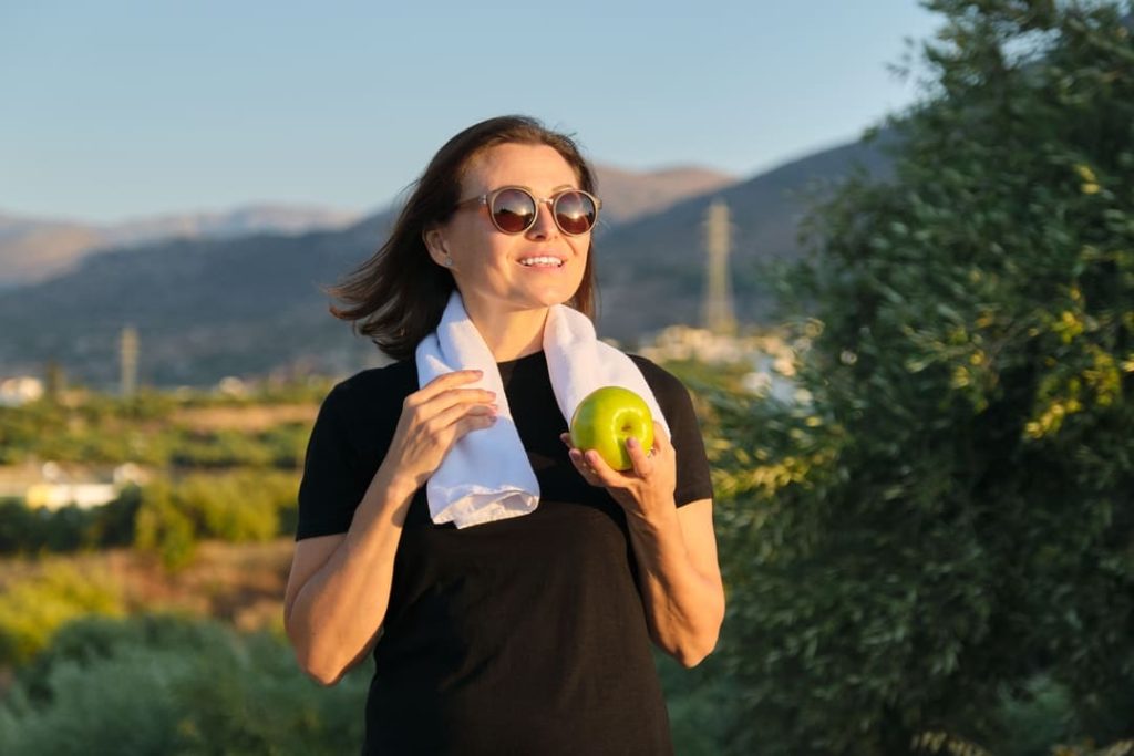 Mulher sorridente ao ar livre com toalha no pescoço e segurando maçã verde, simbolizando hábitos saudáveis.