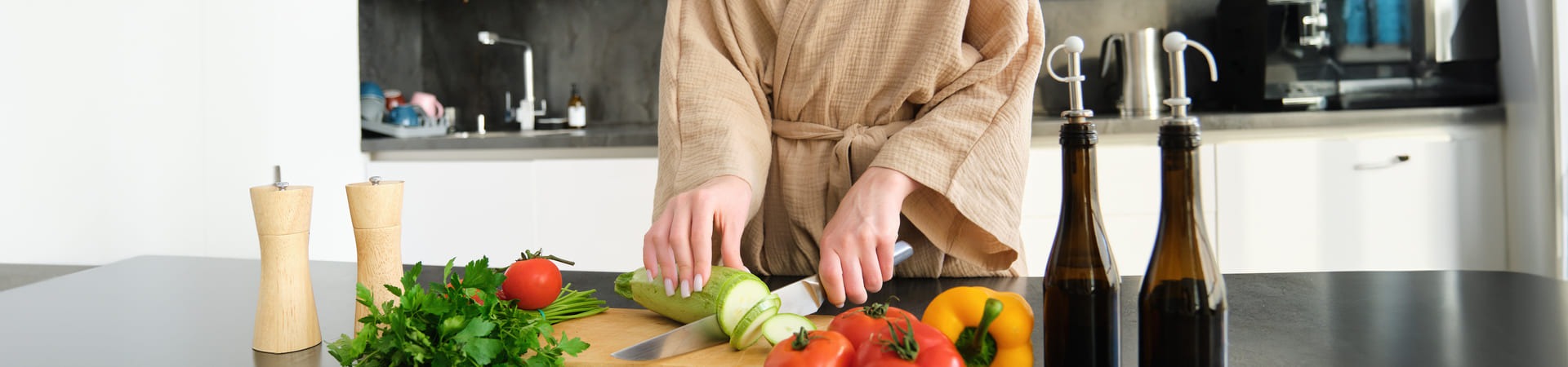Pessoa fatiando um pepino em uma cozinha moderna, cercada por vegetais frescos e temperos naturais sobre a bancada.