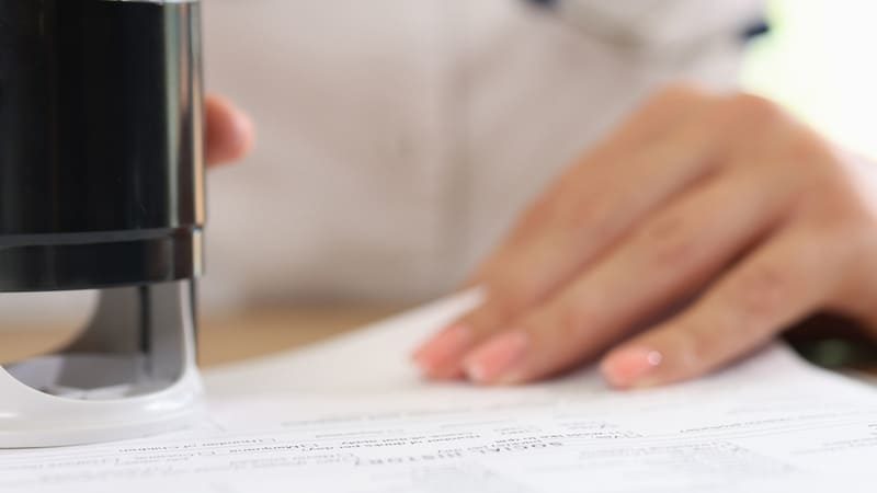 Mão feminina segurando carimbo e autenticando documento sobre uma mesa, possivelmente relacionado a exames de rotina.