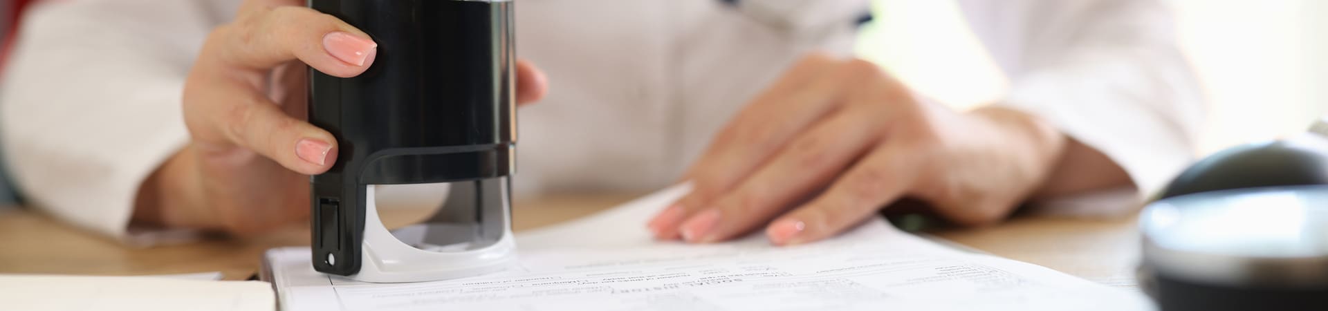 Mão feminina segurando carimbo e autenticando documento sobre uma mesa, possivelmente relacionado a exames de rotina.