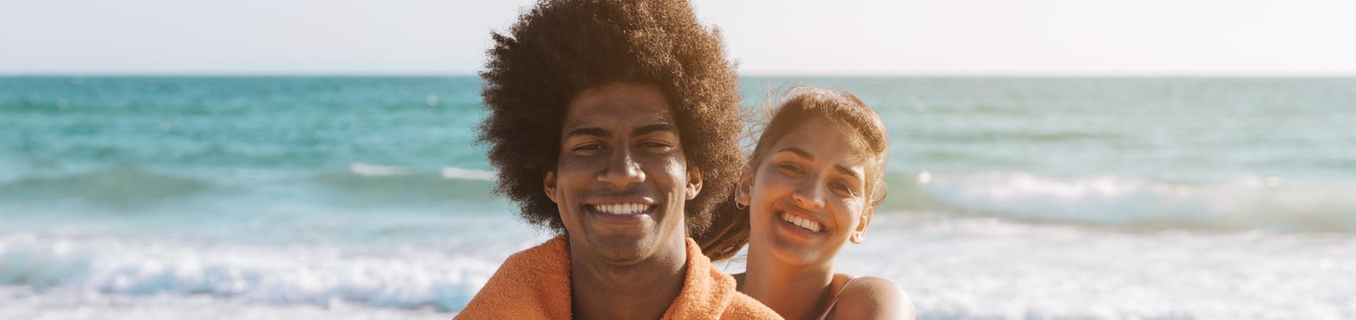 Um casal sorridente está na praia com o mar ao fundo. O homem, com cabelo volumoso, está envolto em uma toalha laranja, e a mulher está ao seu lado, usando uma alça de biquíni. O mar e o céu criam um ambiente relaxante e ensolarado ao fundo.