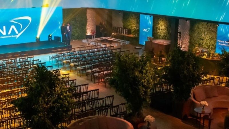 Um grande salão de eventos preparado para uma premiação. O ambiente possui uma decoração sofisticada, com iluminação azul e amarela destacando o palco principal, que exibe o logotipo "ONA" e o texto "Organização Nacional de Acreditação". Há cadeiras organizadas em fileiras à frente do palco e áreas de convivência com sofás e mesas decoradas com arranjos florais. O local conta com paredes cobertas de vegetação e elementos modernos.