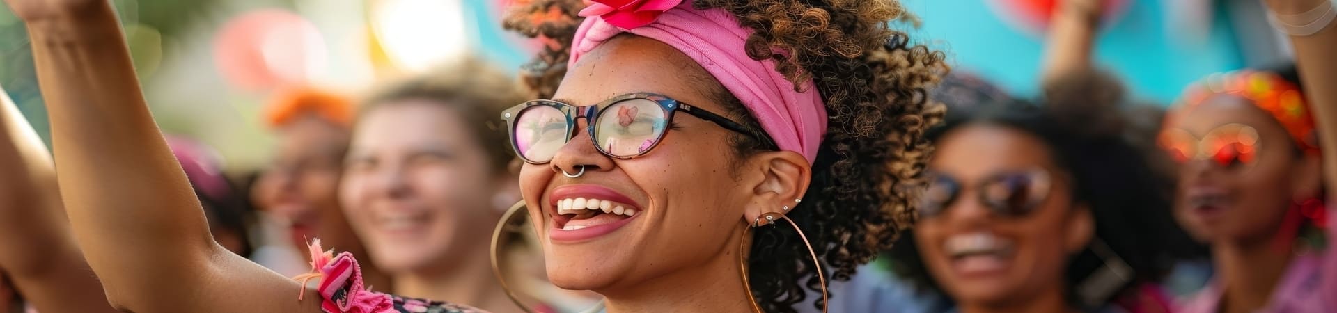 Mulher sorridente em meio a uma multidão animada, usando óculos de grau, um turbante rosa e brincos grandes de argola. O fundo desfocado mostra outras pessoas comemorando, com acessórios coloridos e expressões de alegria.