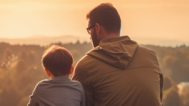 Pai e filho sentados juntos em um mirante, olhando para uma paisagem de montanhas e árvores ao pôr do sol. A imagem transmite um momento de conexão e tranquilidade.