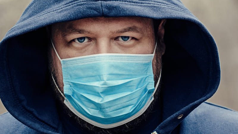 Homem usando capuz azul e máscara cirúrgica azul, olhando diretamente para a câmera.