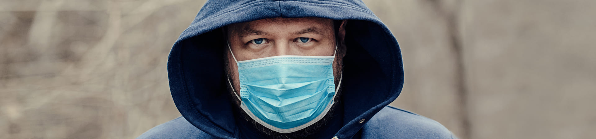 Homem usando capuz azul e máscara cirúrgica azul, olhando diretamente para a câmera.