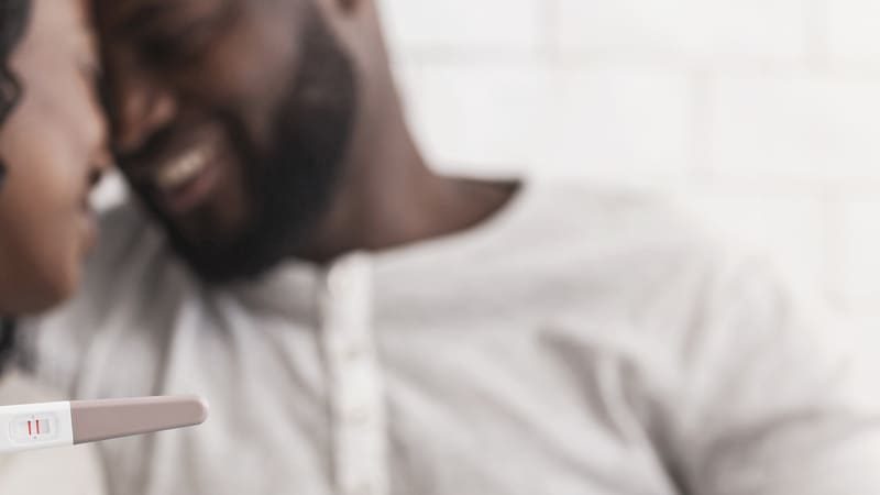 A imagem mostra um casal sorridente e abraçado em um momento de intimidade e alegria. Em primeiro plano, a mulher segura um teste de gravidez com resultado positivo — indicado por duas linhas vermelhas. O foco está no teste, com o casal ao fundo levemente desfocado, olhando um para o outro com expressão de felicidade. O ambiente é iluminado e acolhedor, com uma parede branca de azulejos ao fundo.