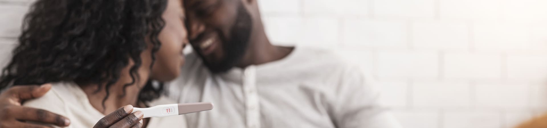 A imagem mostra um casal sorridente e abraçado em um momento de intimidade e alegria. Em primeiro plano, a mulher segura um teste de gravidez com resultado positivo — indicado por duas linhas vermelhas. O foco está no teste, com o casal ao fundo levemente desfocado, olhando um para o outro com expressão de felicidade. O ambiente é iluminado e acolhedor, com uma parede branca de azulejos ao fundo.