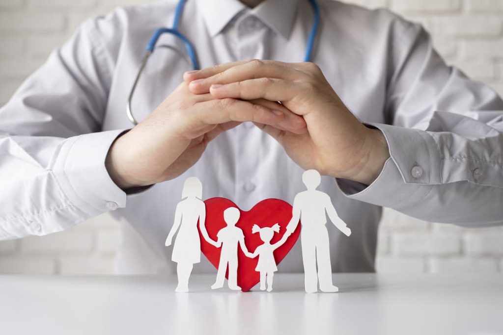 Médico com estetoscópio protegendo com as mãos figuras recortadas de uma família e um coração vermelho ao fundo.
