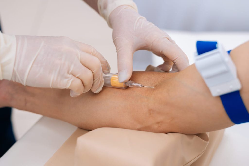 Profissional da saúde coleta sangue do braço de um paciente usando seringa e luvas, representando ações de prevenção e diagnóstico.