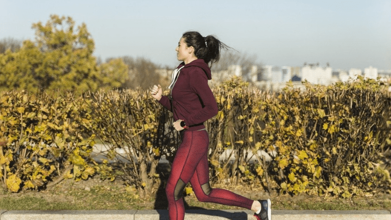 Mulher correndo ao ar livre em um parque, usando roupas esportivas, durante um dia ensolarado. Ela mantém um ritmo constante enquanto passa por arbustos e árvores com folhas de outono.