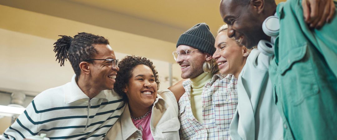 Grupo de cinco pessoas sorrindo e abraçadas, demonstrando amizade e alegria em um ambiente interno iluminado.