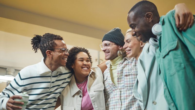 Grupo de cinco pessoas sorrindo e abraçadas, demonstrando amizade e alegria em um ambiente interno iluminado.