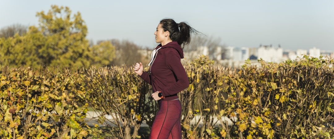 Mulher correndo ao ar livre em um parque, usando roupas esportivas, durante um dia ensolarado. Ela mantém um ritmo constante enquanto passa por arbustos e árvores com folhas de outono.