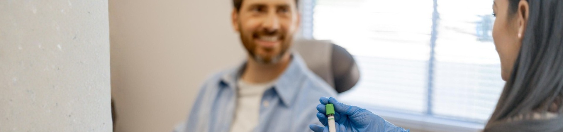 Profissional da saúde com luvas azuis segurando um tubo de coleta de sangue, enquanto um homem sorri sentado na cadeira de atendimento durante a realização de exames laboratoriais.