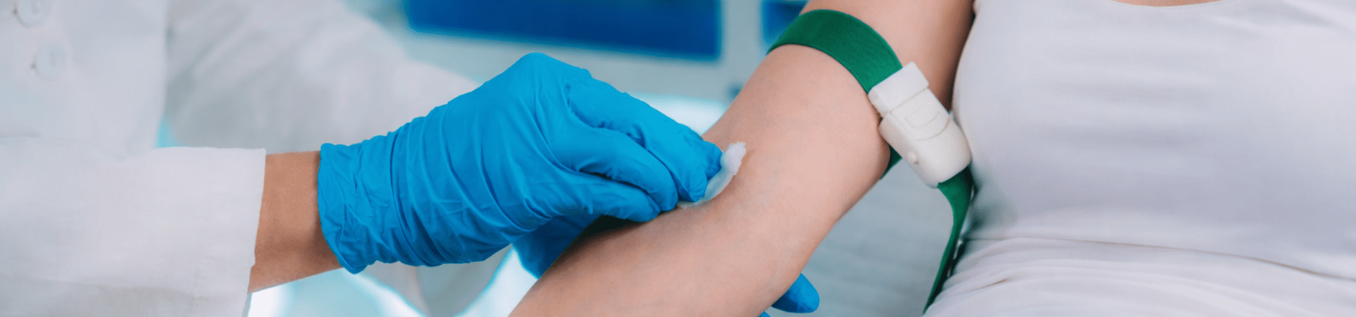 Profissional de saúde usando luvas azuis higieniza o braço de uma pessoa com algodão antes de coleta de sangue, com torniquete verde aplicado.