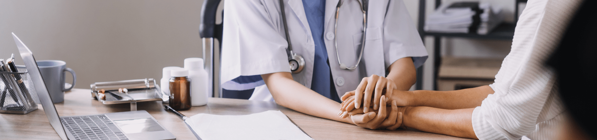 Profissional de saúde segura as mãos de uma paciente sobre a mesa, em gesto de acolhimento durante consulta.
