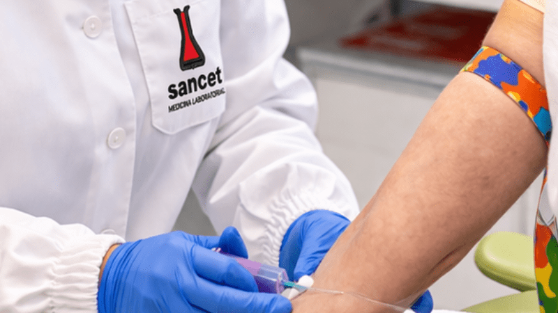 Profissional de saúde usando avental branco com logo Sancet, óculos de proteção e luvas azuis realiza coleta de sangue no braço de uma paciente em ambiente clínico.