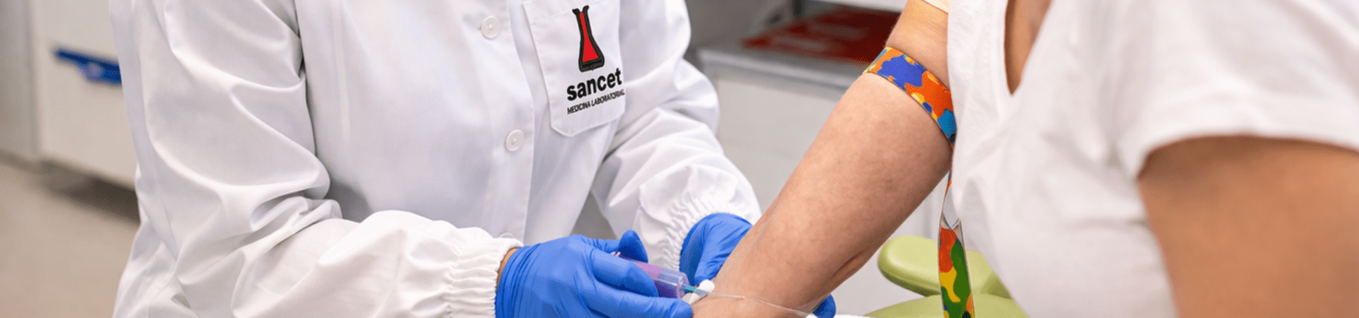 Profissional de saúde usando avental branco com logo Sancet, óculos de proteção e luvas azuis realiza coleta de sangue no braço de uma paciente em ambiente clínico.