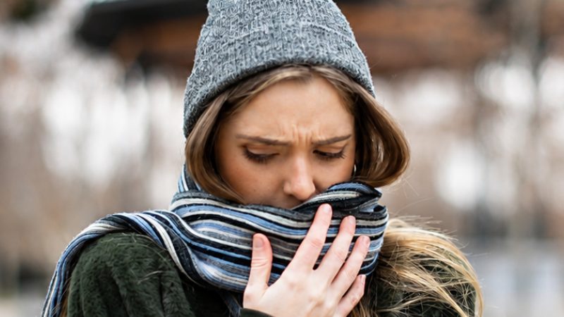 Pessoa agasalhada com gorro e cachecol cobre a boca em um ambiente externo durante o frio, demonstrando desconforto respiratório.
