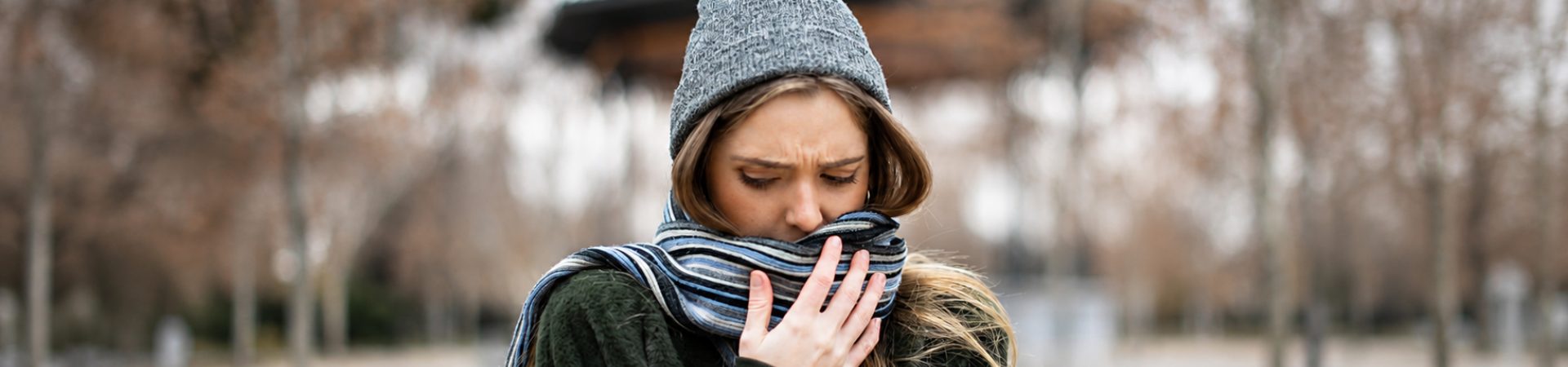 Pessoa agasalhada com gorro e cachecol cobre a boca em um ambiente externo durante o frio, demonstrando desconforto respiratório.